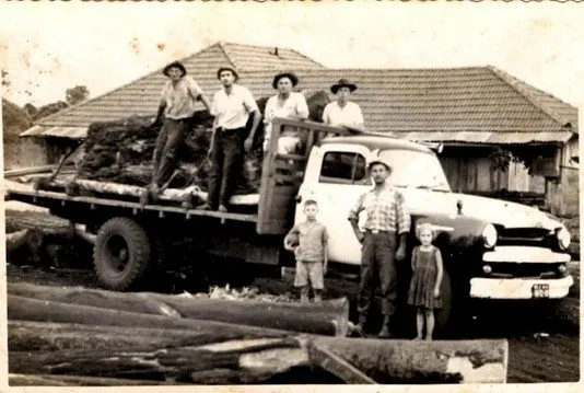 Caminhão antigo carregado de toras, com a equipe e crianças ao lado